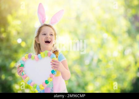 Happy little girl in bunny ears holding a heart board with colorful Easter eggs. Kids celebrate Easter. Children having fun on Easter egg hunt. Stock Photo