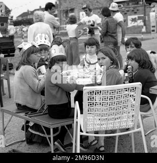 Children in paper hats sit at a table in a street in Crawley, Surrey, and eat ice cream to celebrate Queen Elizabeth II's Silver Jubilee. Stock Photo