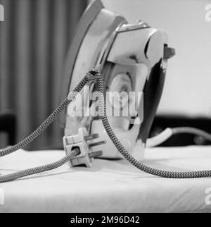 An electric iron with flex and plug, sitting upright on an ironing board.  It has been used so much, the paint has worn off the handle, and the cable is in poor condition. Stock Photo