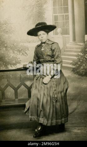 A woman wearing a large hat and pleated skirt, poses on a bench in a photographic studio Stock Photo