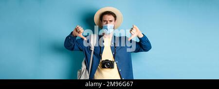 Covid-19 and summer holidays concept. Disappointed tourist in medicla mask frowning, showing thumbs down, negative feedback about travel, standing on Stock Photo