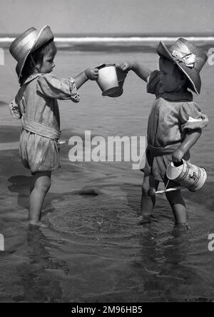 Two toddlers paddling in the sea, each one with a bucket in hand.  The one on the right also carries a small watering can. Stock Photo