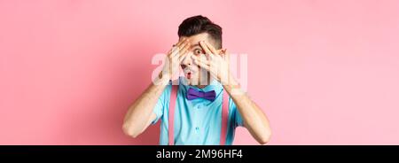 Intrigued funny guy in bow-tie, peeking through fingers at something interesting, checking out something cool, standing on pink background Stock Photo