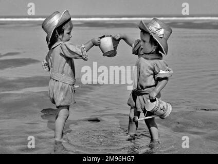 Two toddlers paddling in the sea, each one with a bucket in hand. The one on the right also carries a small watering can. Stock Photo