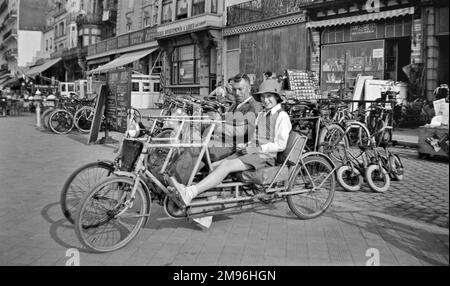 A couple on a four-wheeled two-seater cycle (also known as a velo double) outside the Taverne Emile in a town in Belgium. Stock Photo