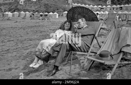 A man and a woman sitting in deckchairs on the beach at Newquay, Cornwall, with an oriental-style parasol to shade them from the sun. Stock Photo