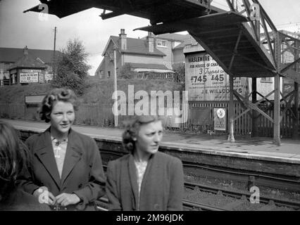 Two young women at a railway station in South London.  An advertisement on the opposite platform is for property, houses of character for £745 freehold.  Another sign advertises self-contained flats to let. Stock Photo