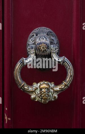 Closeup view of traditional vintage brass door knocker with face of man isolated on dark red wooden door, Montpellier, France Stock Photo