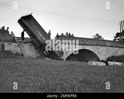 A lorry (Lewin's Lorries for Loads) which has fallen off a low bridge onto a grassy area.  People stand around looking fairly helpless. Stock Photo