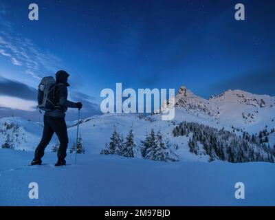 Night photo climber stands on top of a mountain in the snow and looks at the surrounding mountains with starry sky Stock Photo