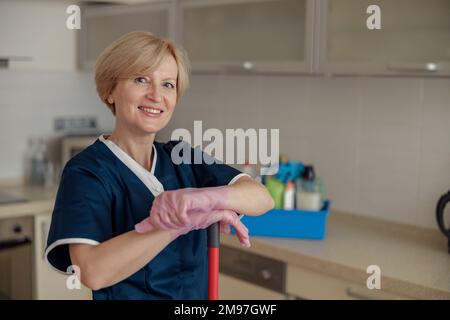 Portrait of smiling senior housekeeping standing with mop on kitchen and looking at camera Stock Photo