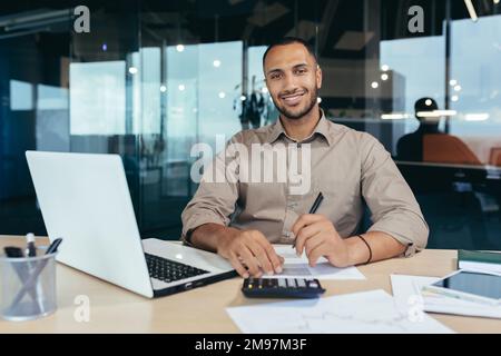 Portrait of young successful hispanic businessman inside office, man smiling and looking at camera, paper worker happy with achievement results sitting at workplace with laptop. Stock Photo