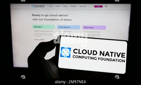 Person holding cellphone with logo of Cloud Native Computing Foundation (CNCF) on screen in front of webpage. Focus on phone display. Stock Photo