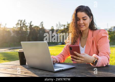 Businesswoman sitting at a table in a park, outdoors working on her laptop while checking her cell phone. Stock Photo