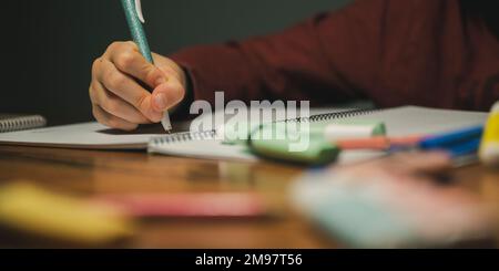 Low angle closeup view of a child making homework, writing in his notebook. Marker pens an pencils lying on a desk. Stock Photo