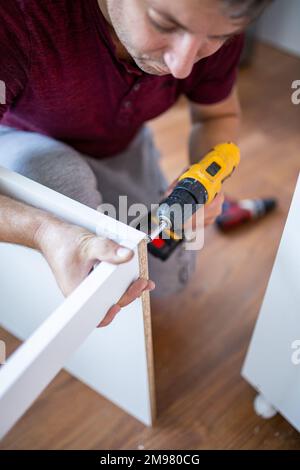 Furniture Installation. Man Using Electric Drill Assembling And Fixing Wooden Shelf Furnishing Room At Home. Stock Photo