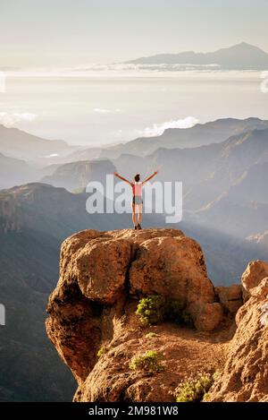 Rear view of a woman standing on rocks with Roque Nublo natural park and Mount Teide in the background, Gran Canaria, Canary Islands, Spain Stock Photo