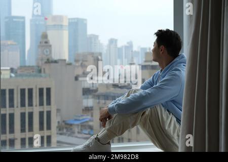 lonely Chinese young man sit at window sill, look out window to see urban city buildings at Shanghai, China Stock Photo