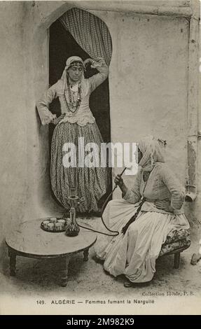 Two women smoke from a hookah pipe in Algeria, North Africa. One woman is seated and holds the stem of the sheesha pipe near her mouth, ready to inhale. The other woman stands in a doorway near her, watching and waiting for her turn. Both females exhibit traditional dress and headscarves typical of the region. Stock Photo