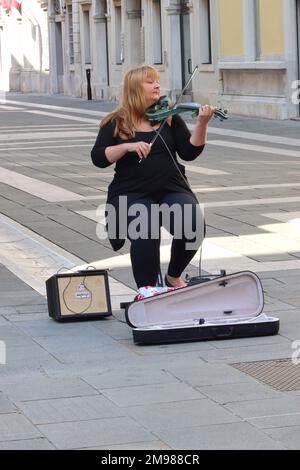 A street musician plays a Stagg 4/4 Electric Violin connected to an amplifier, in the pedestrianised Via Saint Nicolo, Trieste, Italy. Stock Photo