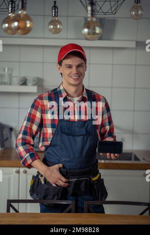 Smiling pleased repair technician standing at the customer kitchen and holding his cellphone in the hand Stock Photo