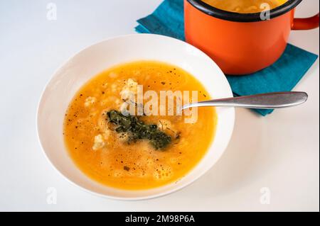 Carrot soup with pasta and fried rucola in white plate, blue towel and part of orange pot on white background. Stock Photo