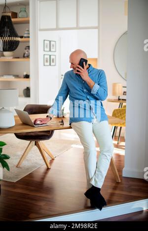 Full length of happy middle aged man making a call and using laptop while working from home. Confident male standing at desk in the living room. Home Stock Photo