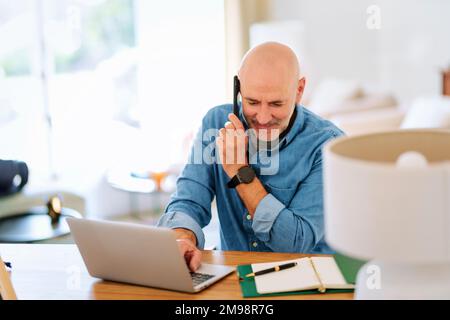 Happy middle aged man making a call and using laptop while working from home. Confident male sitting in the living room and having a call. Home office Stock Photo