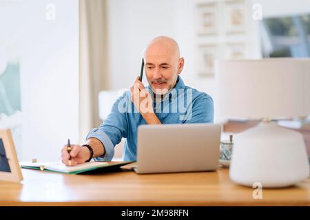 Happy middle aged man making a call and using laptop while working from home. Confident male sitting in the living room and having a call. Home office Stock Photo