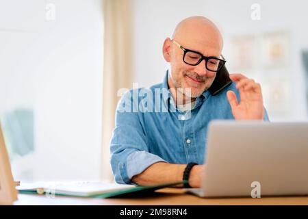Happy middle aged man making a call and using laptop while working from home. Confident male sitting in the living room and having a call. Home office Stock Photo