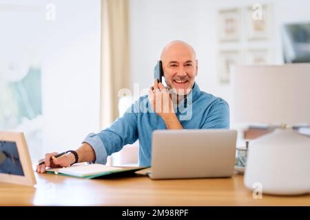 Happy middle aged man making a call and using laptop while working from home. Confident male sitting in the living room and having a call. Home office Stock Photo