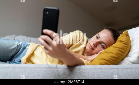 Woman with blank expression face lying on sofa and using her cell phone. Stock Photo