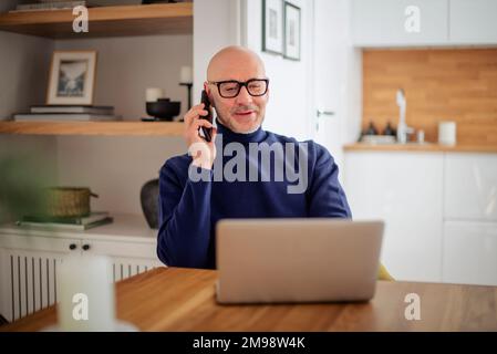 Confident middle aged man making a call and using laptop while working from home. Confident male sitting in the kitchen and having a call. Home office Stock Photo