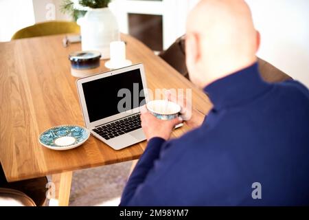 Overhead shot of a bald man using laptop while working from home. Blank screen. Confident male drinking tea and sitting at table. Home office. Stock Photo