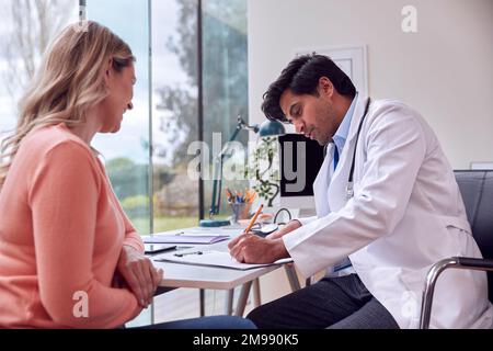 Male Doctor Or GP Meeting Mature Female Patient For Appointment In Office Making Notes On Clipboard Stock Photo