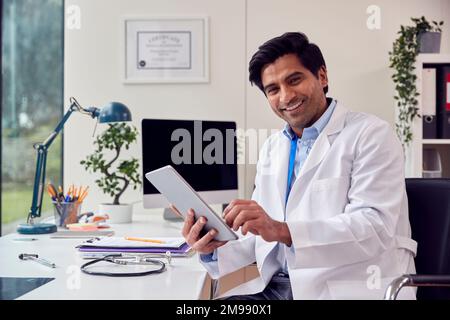 Portrait Of Male Doctor Or GP Wearing White Coat Sitting At Desk In Office Using Digital Tablet Stock Photo