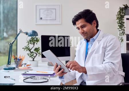 Male Doctor Or GP Wearing White Coat Sitting At Desk In Office Using Digital Tablet Stock Photo