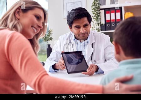 Doctor Or GP In White Coat Meeting Mother And Son For Appointment Looking At Scan On Digital Tablet Stock Photo