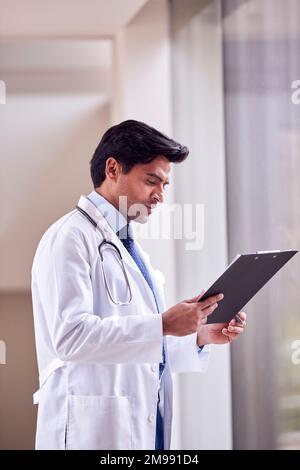 Male Doctor Wearing White Coat Standing In Hospital Corridor Looking At Clipboard Stock Photo