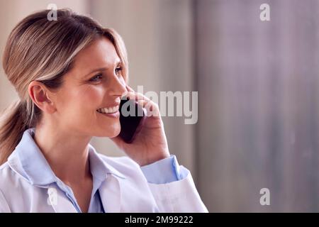 Female Doctor Wearing White Coat Standing In Hospital Corridor Talking On Mobile Phone Stock Photo