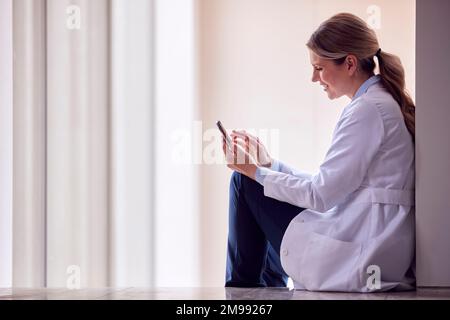 Female Doctor Wearing White Coat Sitting On Floor In Hospital Corridor Using Mobile Phone Stock Photo
