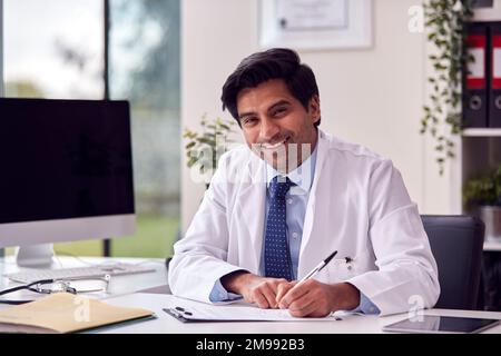 Portrait Of Male Doctor Or GP Wearing White Coat At Desk In Office Writing Notes On Clipboard Stock Photo