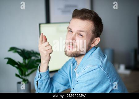Agree or you're right gesture: cute bearded portrait of caucasian male freelance software engineer sitting at working place in office looking at camera with computer on background. High quality image Stock Photo