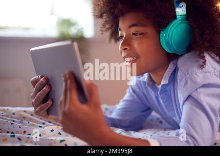 Boy In Bedroom Lying On Bed Wearing Headphones Using Digital Tablet Stock Photo