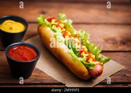 Hot dog with mustard, ketchup and green salad in bun, served with potato chips on a rustic wooden board. Fast food and delicious. Stock Photo