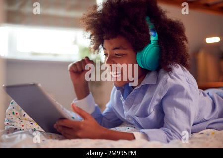 Boy In Bedroom Lying On Bed Wearing Headphones Using Digital Tablet Stock Photo