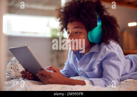 Boy In Bedroom Lying On Bed Wearing Headphones Using Digital Tablet Stock Photo