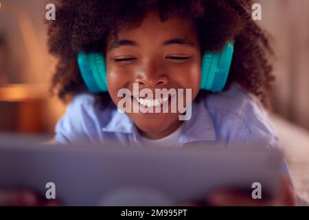 Boy In Bedroom Lying On Bed Wearing Headphones Using Digital Tablet Stock Photo