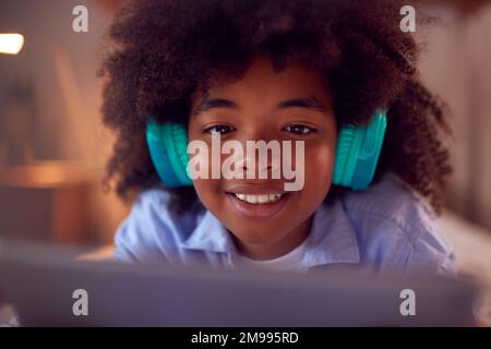 Boy In Bedroom Lying On Bed Wearing Headphones Using Digital Tablet Stock Photo