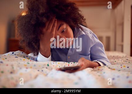 Worried Boy In Bedroom Lying On Bed Holding Mobile Phone Being Bullied Online Stock Photo